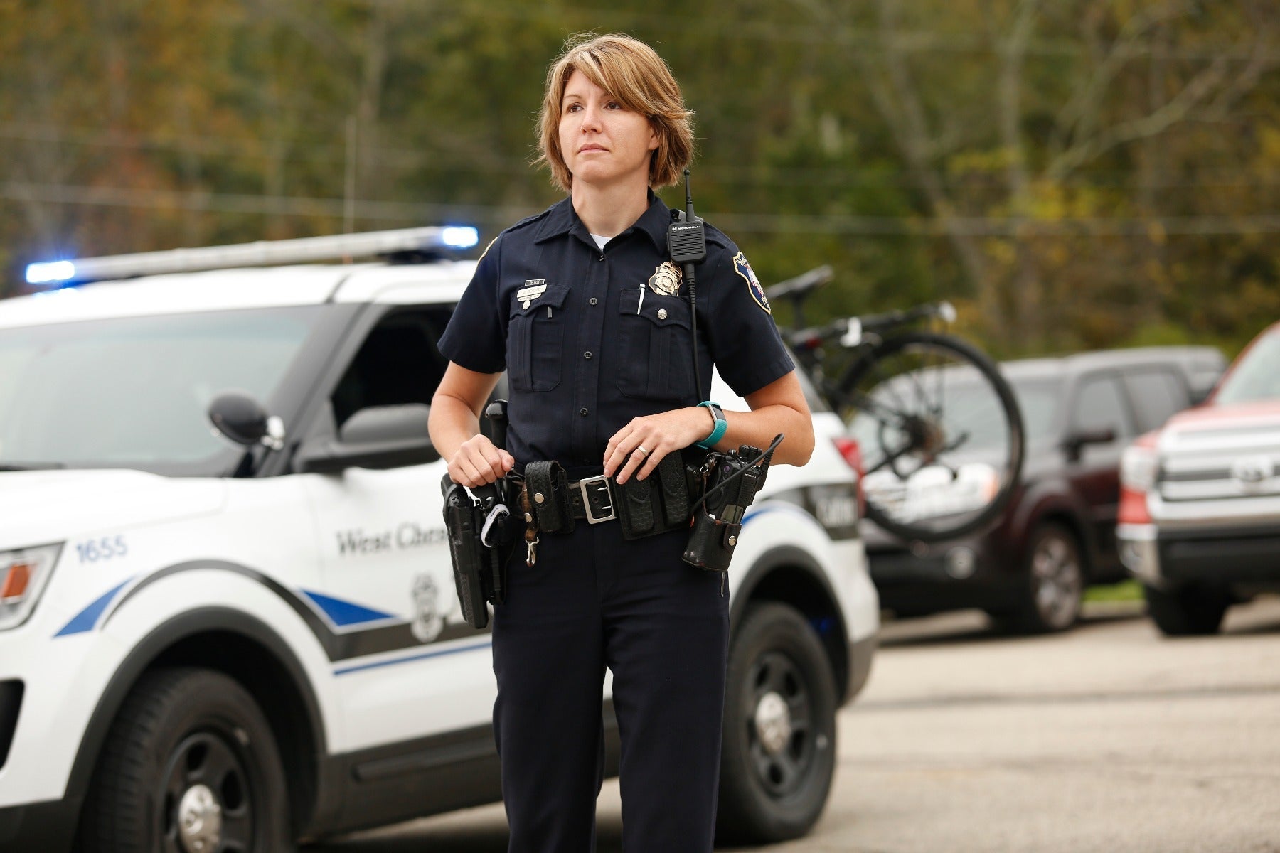 Photograph West Chester Police officer in uniform standing near a police car and bicycle with a teal fitness tracker and a silver badge visible a West Chester Police vehicle number 1655 in the background