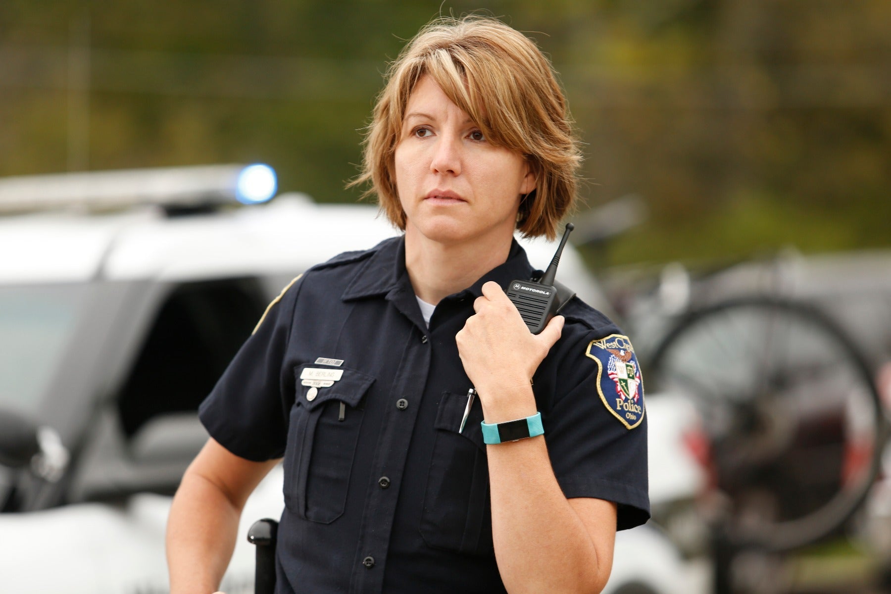 Photograph of a female West Chester Police officer in Ohio holding a Motorola radio with a teal fitness tracker and a silver badge near a police car with flashing lights and a blurred bicycle in the background