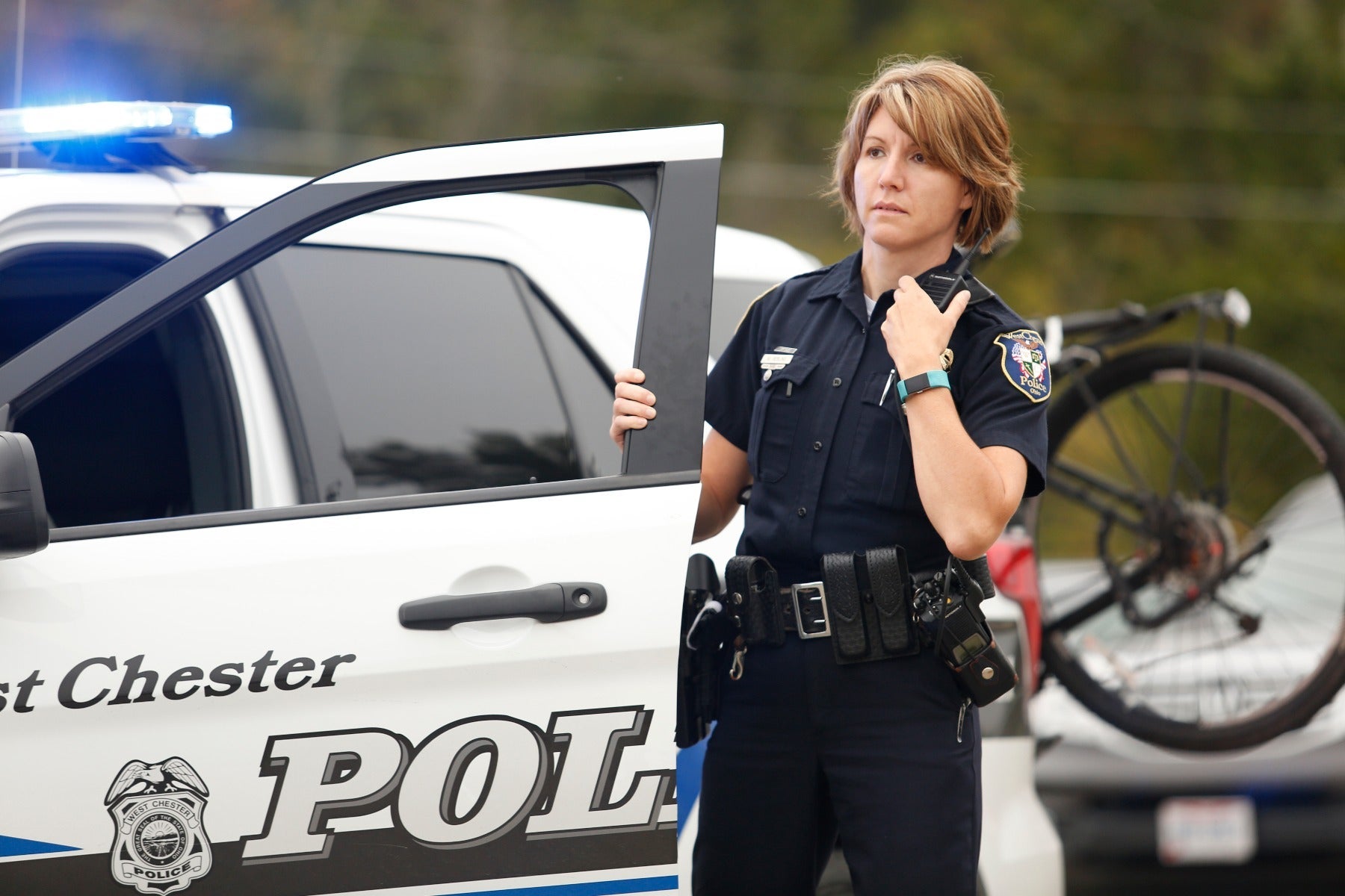 Photograph A female West Chester Police officer in uniform using a radio by her patrol car with blue flashing lights and a bicycle visible in the background a teal fitness tracker and a police badge are also visible