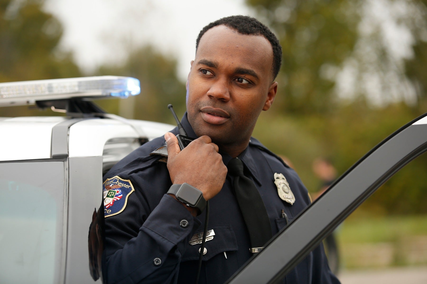 Photograph of a police officer using a radio next to his patrol car, showcasing a West Chester Police Department badge, a smart watch, and a blue flashing light on the vehicle