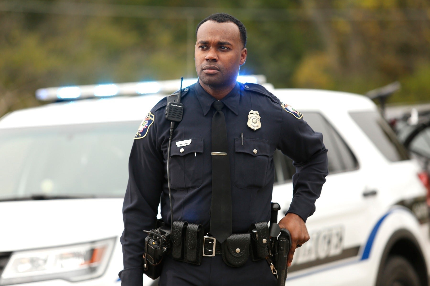 Photograph of a police officer in a dark uniform with a police badge and Motorola radio standing in front of a police car with flashing blue lights and a West Valley Police decal