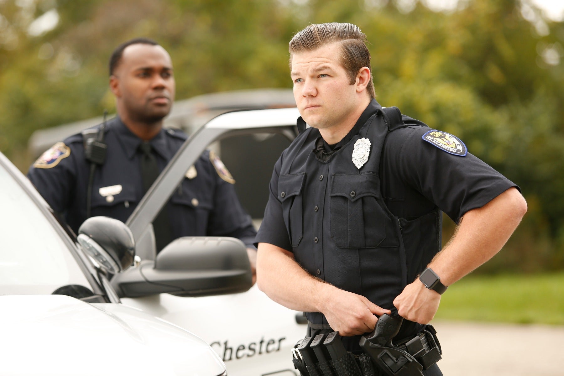 Photograph of two police officers in uniform standing by a Chester County Police car featuring a silver badge, a black Apple Watch, and a dark handgun in a holster.