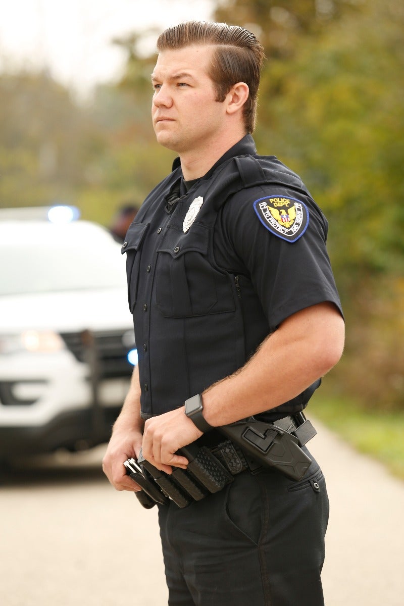 Photograph of a police officer in uniform standing outdoors near a police vehicle showcasing a dark uniform, a distinctive police department patch, and a black smartwatch.