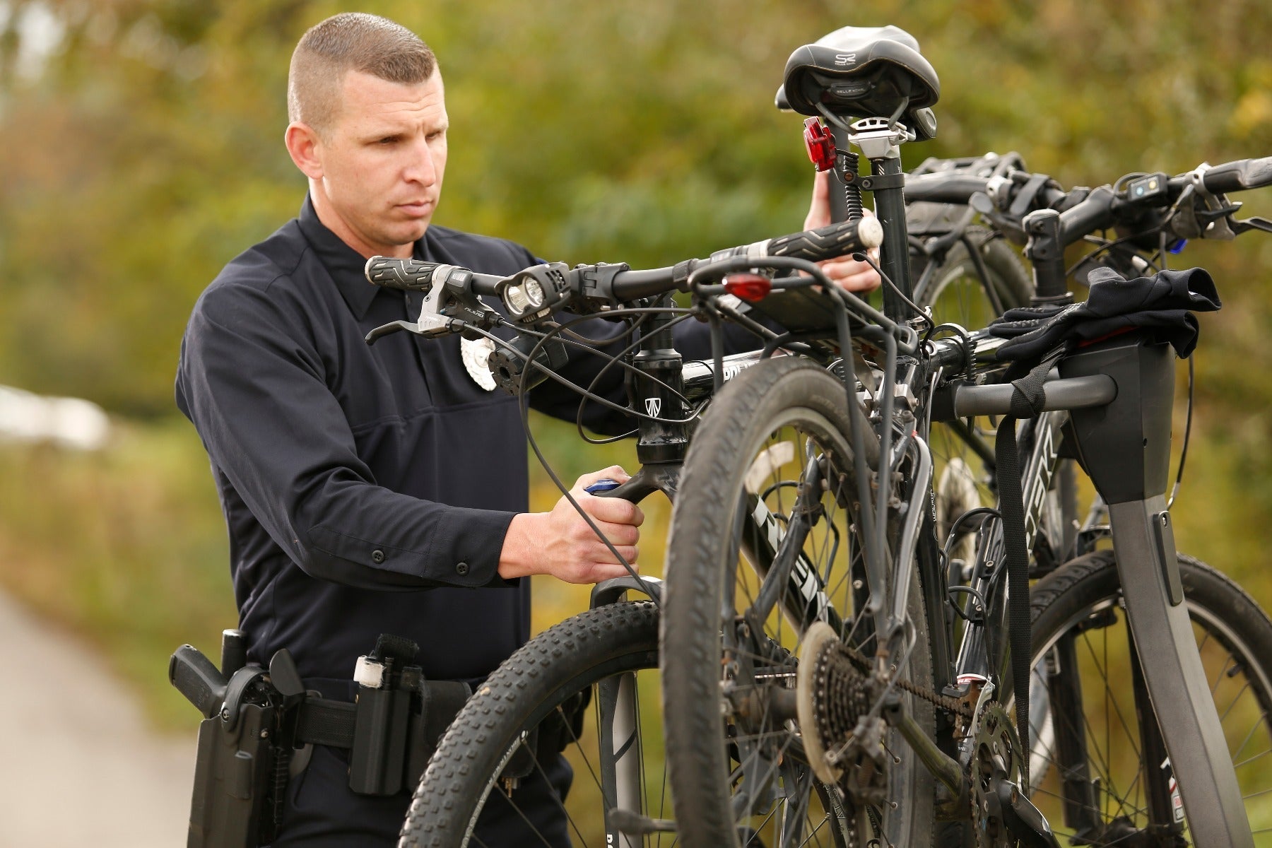 Photograph A police officer prepares a mountain bike with a black frame and red rear light in a park-like setting with autumnal foliage