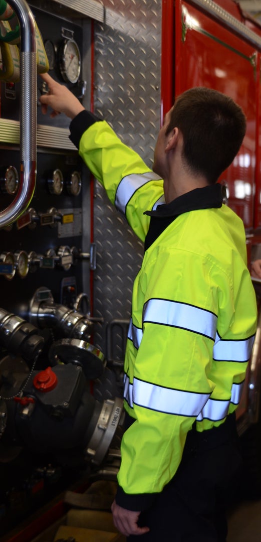 Photograph shows a firefighter in a bright lime green jacket with reflective stripes inspecting the gauges and hoses of a red firetruck with diamond plate metal siding
