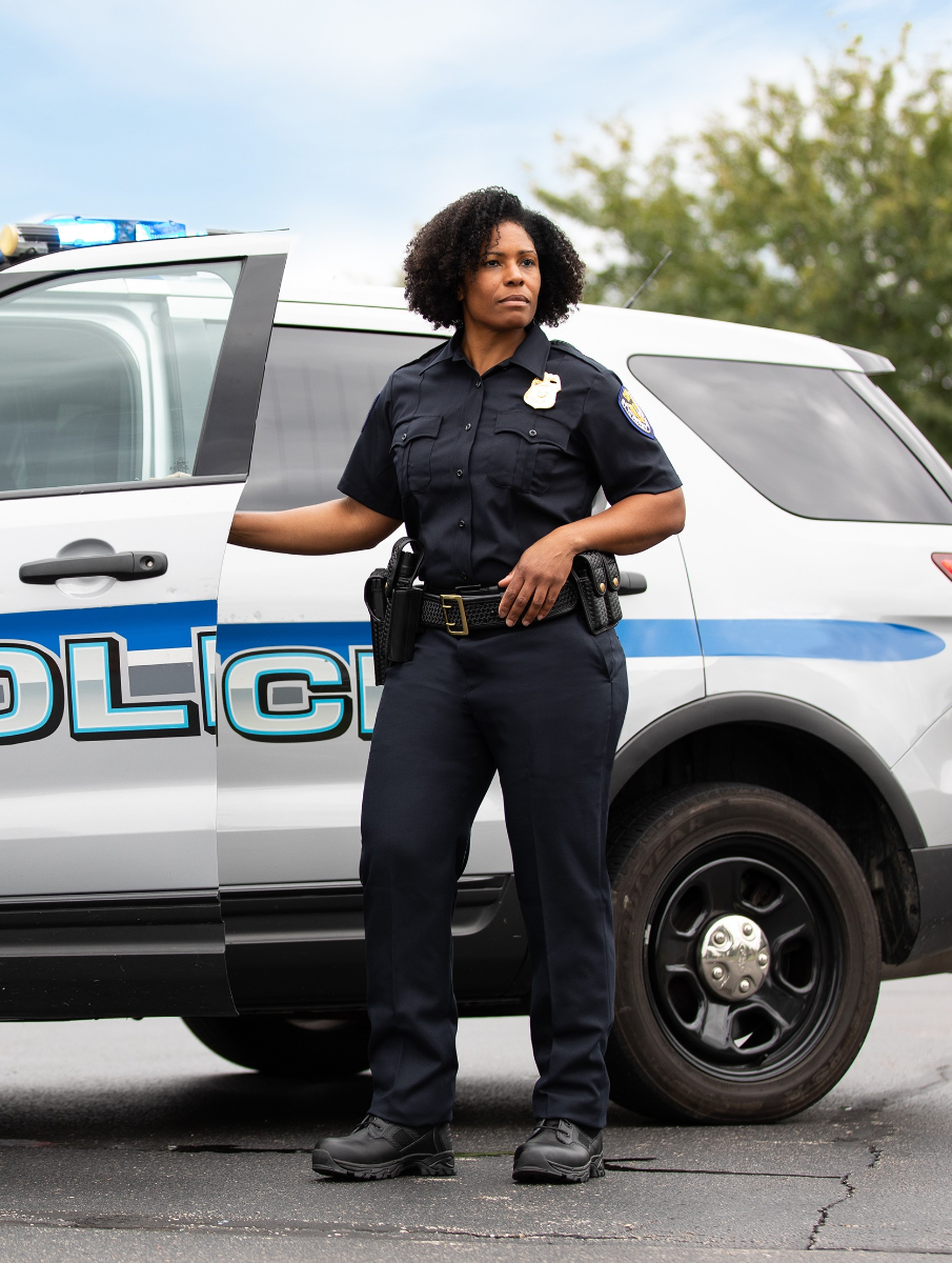 Photograph of a Black female police officer standing by her police car with blue and white POLICE lettering, a serious expression, sturdy black boots, and a dark uniform with a badge outdoors near green trees.