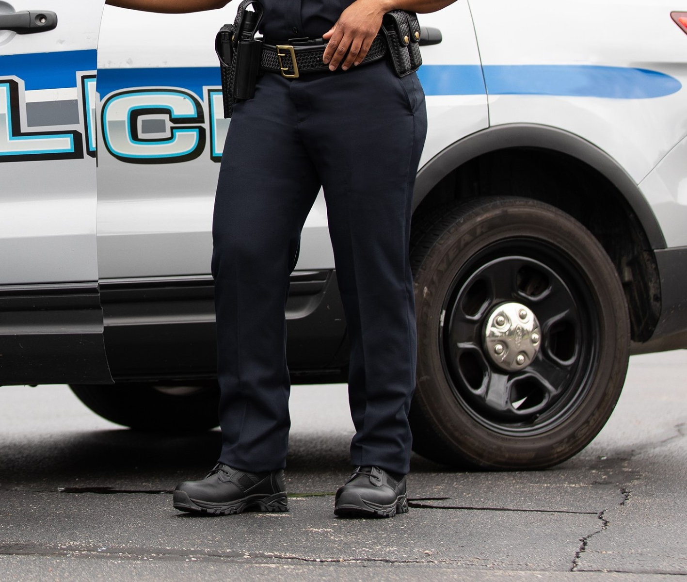 Photograph Police officer standing next to a police vehicle with a blue and gray design, black tactical boots, and a gun belt, near a cracked asphalt surface.