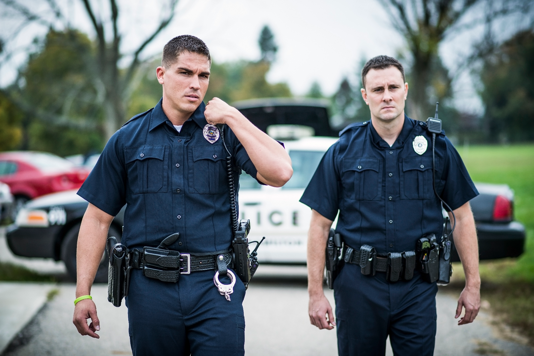 Photograph two police officers in dark blue uniforms with silver badges and utility belts standing near a police car and damaged red car in a park-like setting with green grass and trees in the background