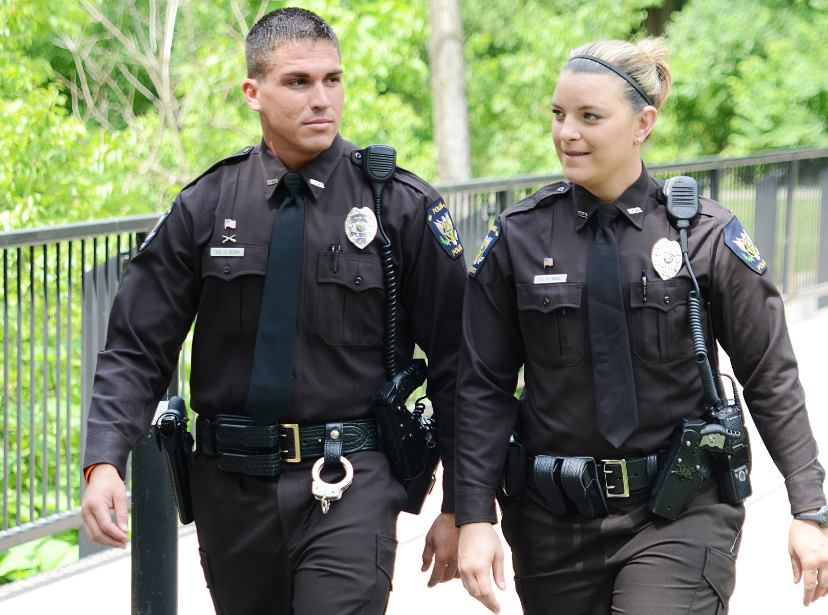 Photograph Two police officers in dark uniforms with gold buckles and name tags walk near a black metal fence and lush green trees