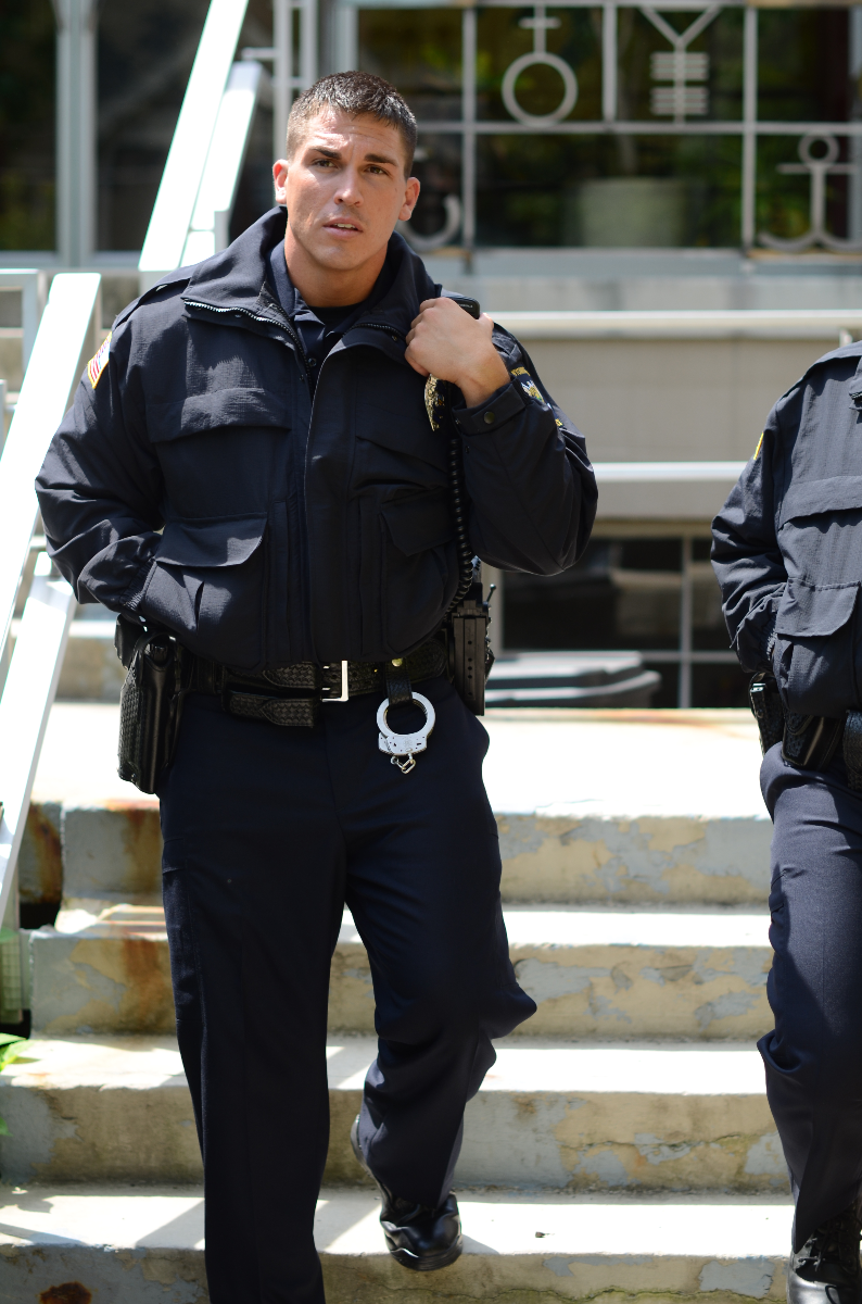 Photograph Police officer on steps outside building with symbolic window details and US flag patch on dark uniform shiny handcuffs