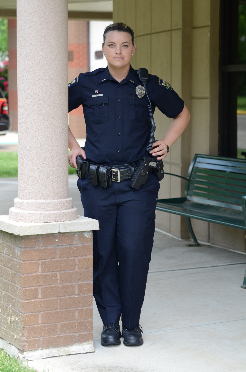 Photograph Police officer standing outside a brick building near a green bench wearing a dark blue uniform with a gold belt and black shoes