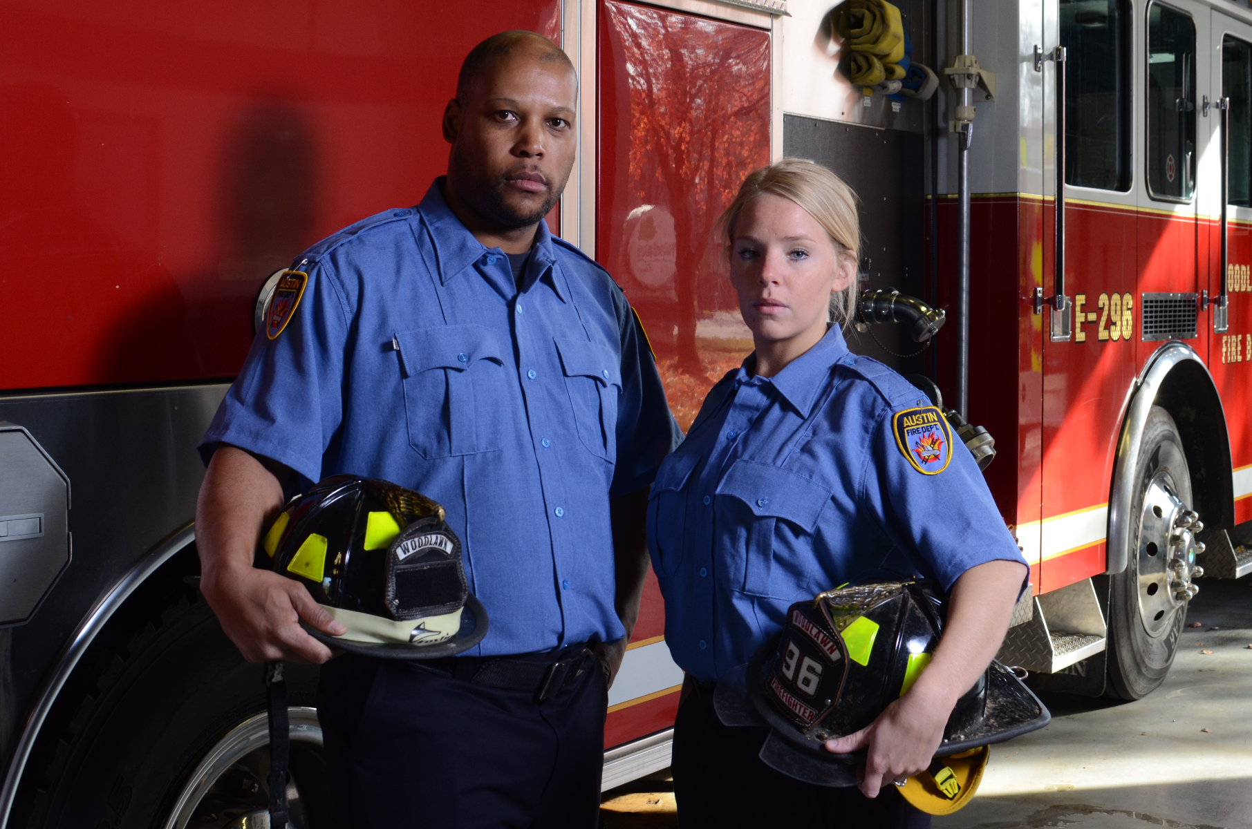 Photograph of two firefighters, a man and a woman, in blue uniforms standing in a fire station next to a bright red firetruck E-296 with their helmets, featuring yellow reflective accents and numbered patches