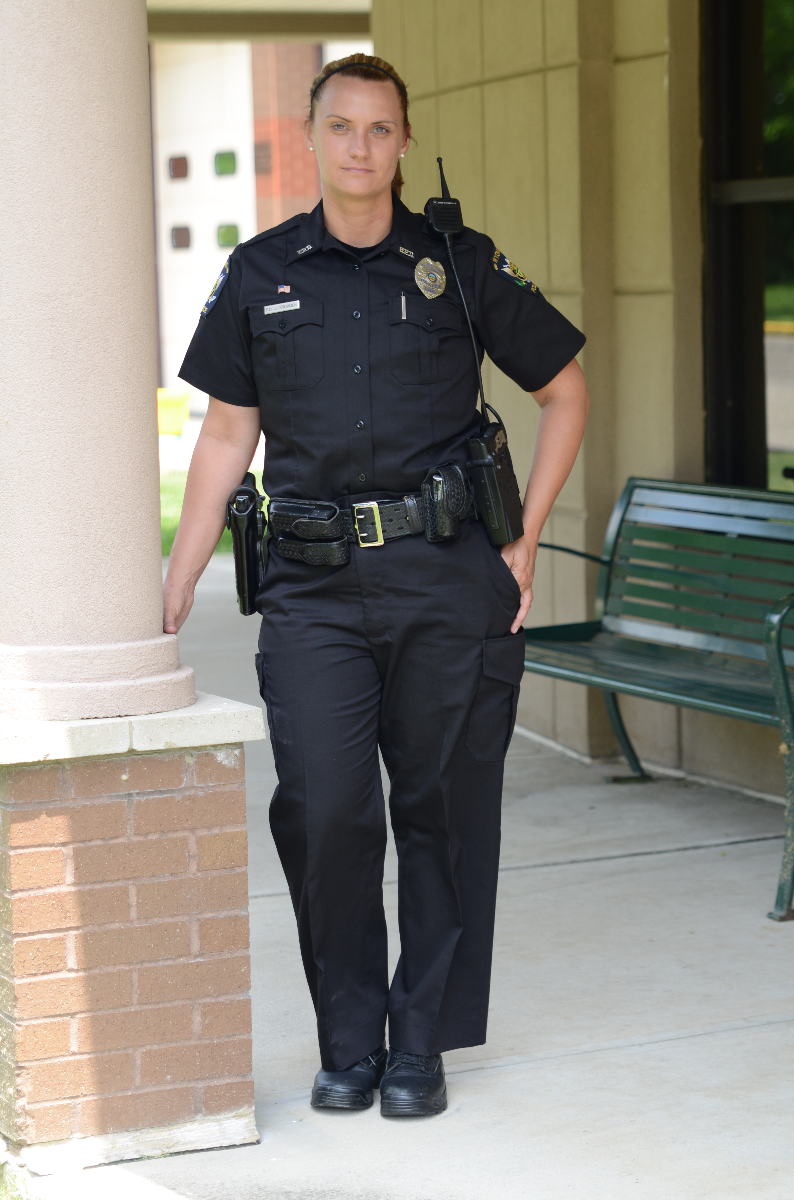 Photograph of a female police officer in uniform standing near a brick pillar and a green bench outside a building with visible windows and a tan wall