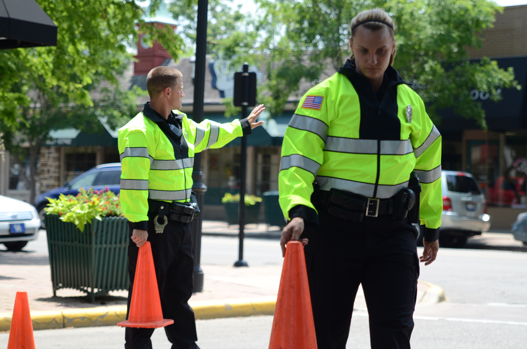 Photograph of two police officers in bright neon-yellow jackets directing traffic with orange cones near a quaint town square featuring a red brick building and lush green trees