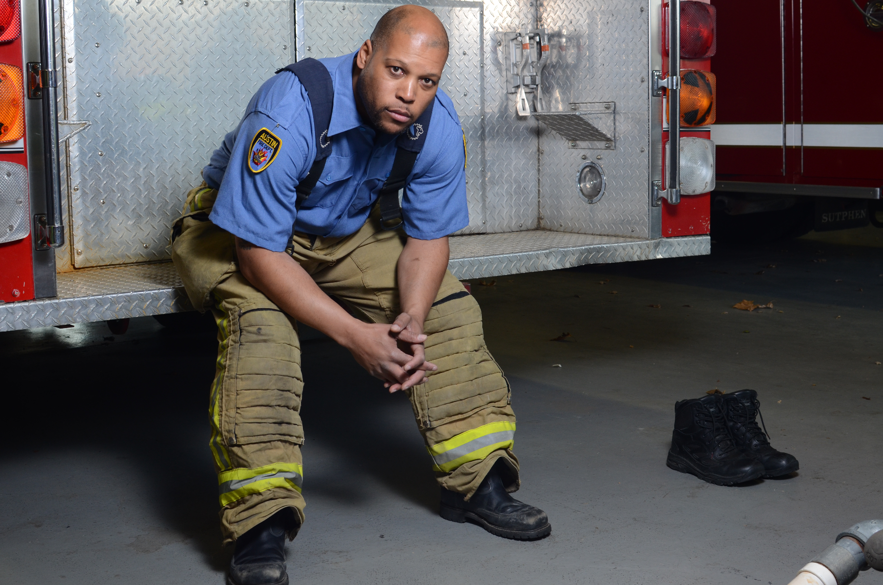 Photography Austin Fire Department firefighter seated on firetruck in fire station wearing soiled tan fire pants blue shirt and black boots near his black work boots with reflective yellow accents and a red firetruck in the background
