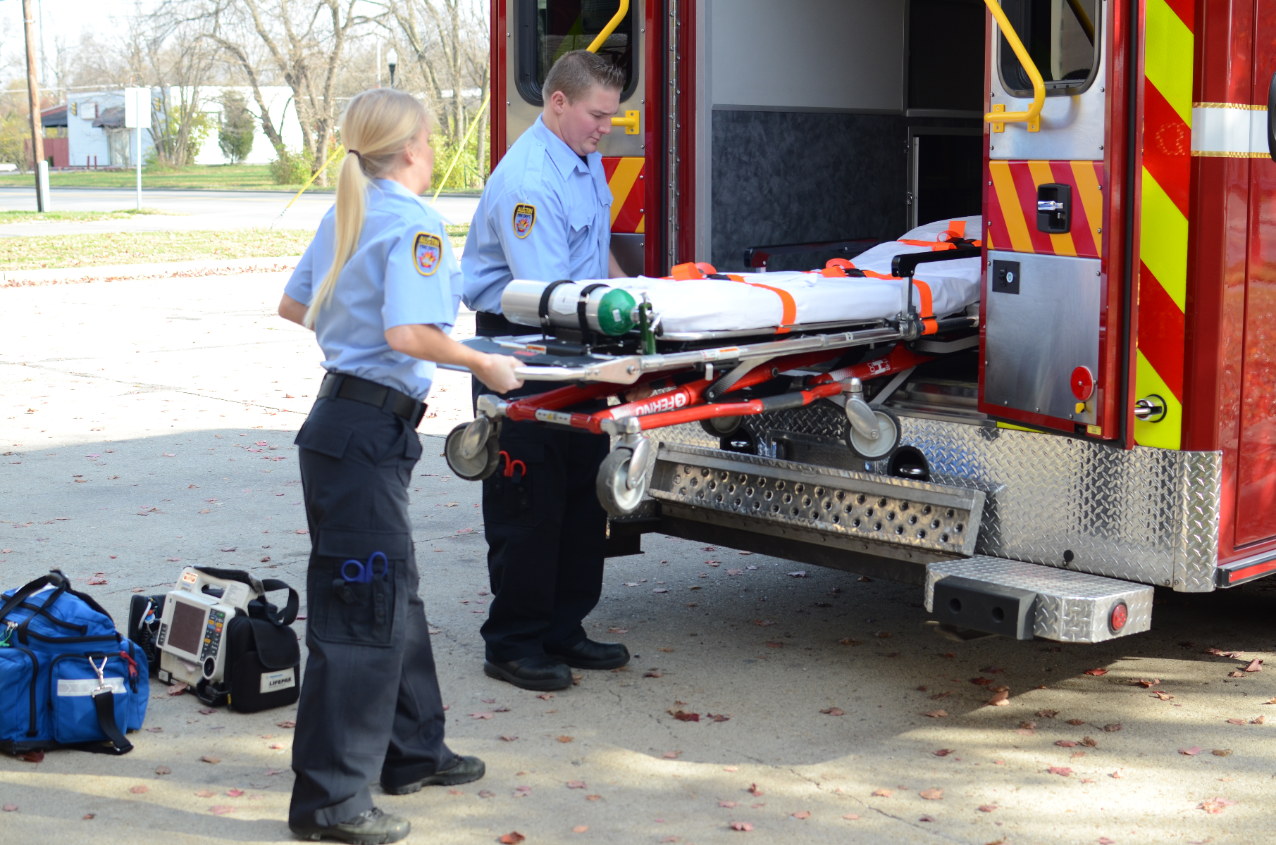 Photograph paramedics loading a stretcher into an ambulance near a residential area showing a red and yellow striped ambulance a bright blue uniform and a green oxygen tank