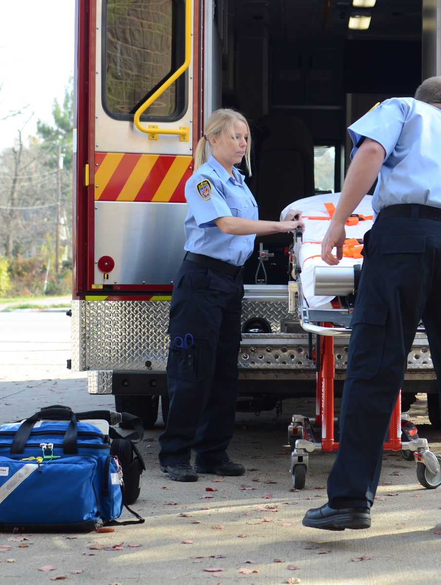 Photograph of two EMTs near an ambulance loading a stretcher showing a blue uniform a red and yellow striped ambulance and scattered autumn leaves
