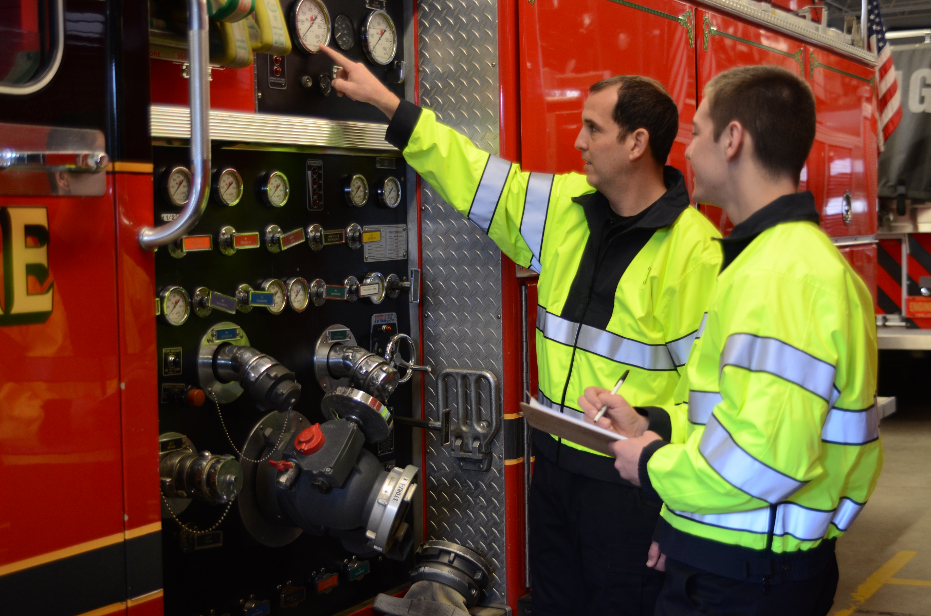 Photograph showing two firefighters in bright yellow jackets inspecting a fire truck's control panel featuring numerous gauges and valves in a fire station with a partially visible American flag and a red fire truck in the background featuring metallic silver accents and a black stripe