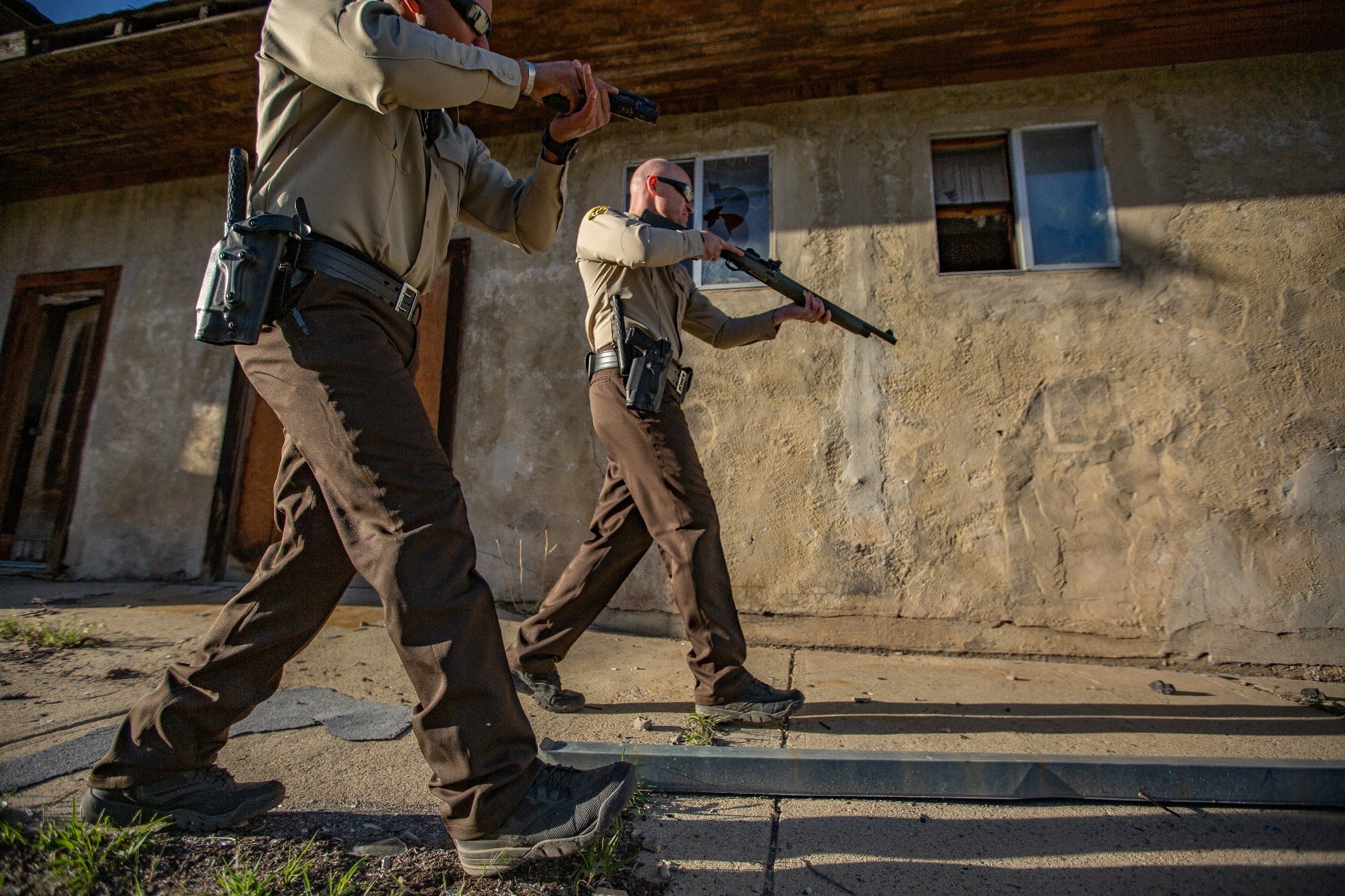 Photograph showing two law enforcement officers in brown uniforms tactical maneuvering near a dilapidated building with a broken window and weathered tan stucco walls carrying handguns and a long gun their dark tactical boots and gear contrasting sharply with the dusty setting