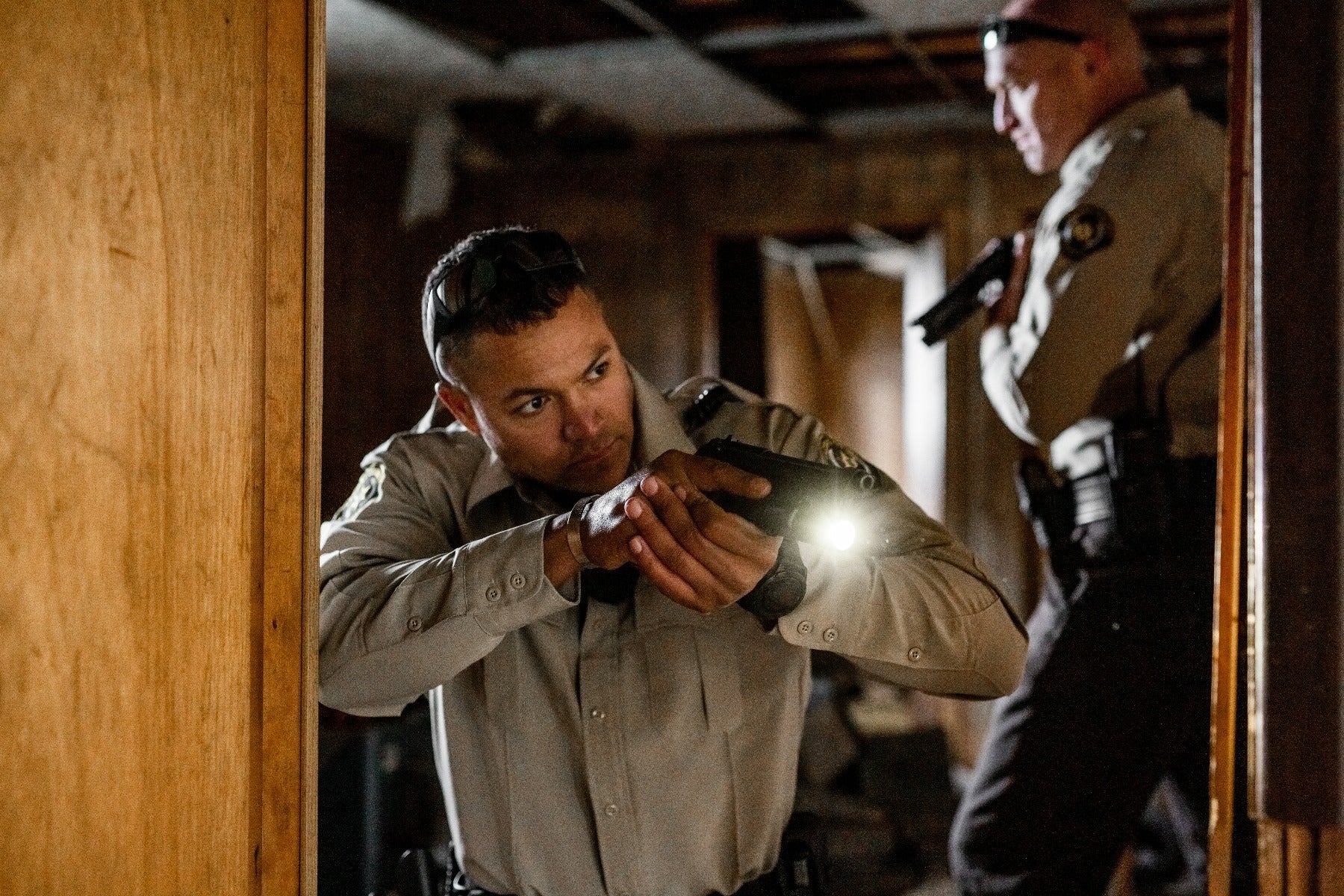 Photograph depicting two law enforcement officers in a dimly lit interior, one aiming a handgun with attached flashlight at a doorway, the other holding a shotgun, showcasing the officers’ tactical gear, a weathered wooden door, and the beam of light cutting through the darkness.