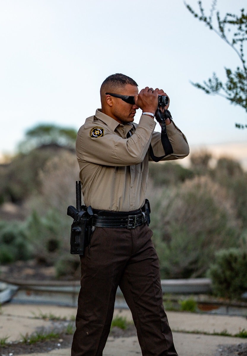 Photograph a uniformed law enforcement officer using binoculars outdoors in a desert environment with brown pants, a tan shirt and a sheriff's badge visible