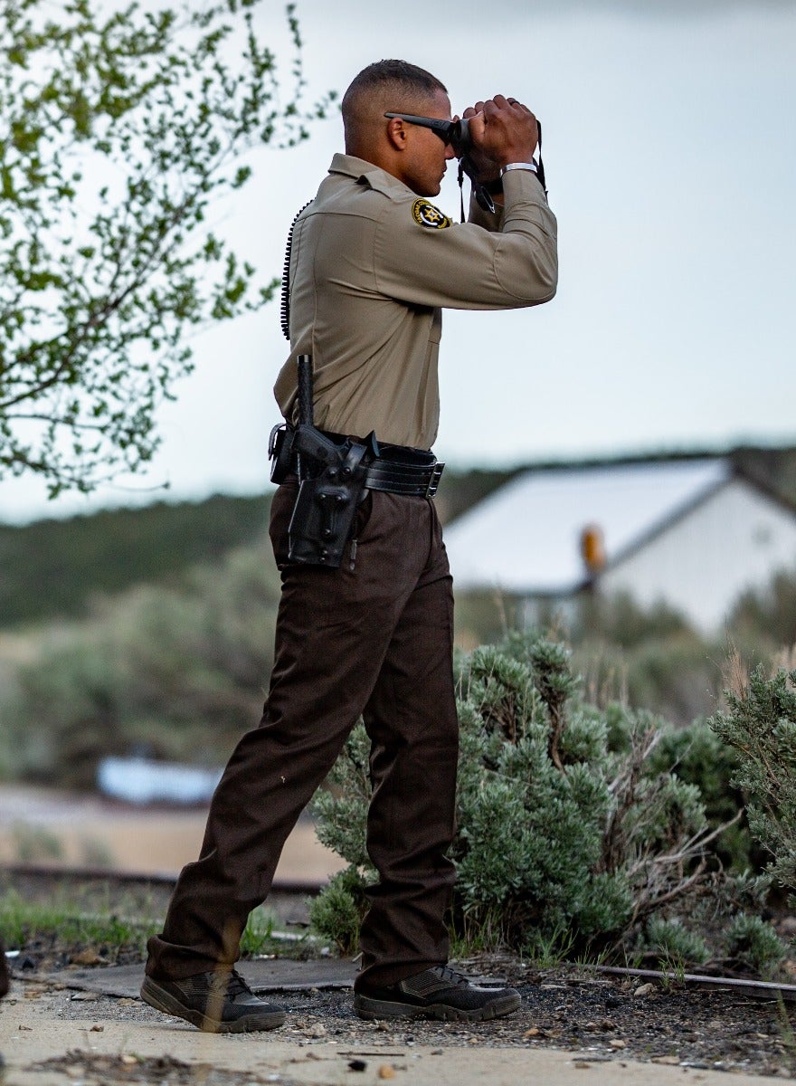 Photograph of a law enforcement officer in tan uniform and brown pants using binoculars near a weathered building and train tracks featuring sagebrush and a dark-colored holster