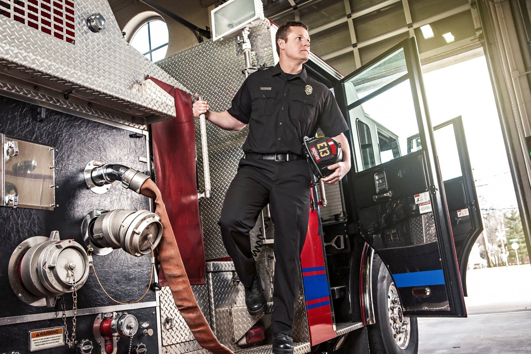 Photograph of a firefighter standing by a firetruck in a fire station featuring a diamond-plate metal exterior, red and blue stripes, and a tan fire hose.