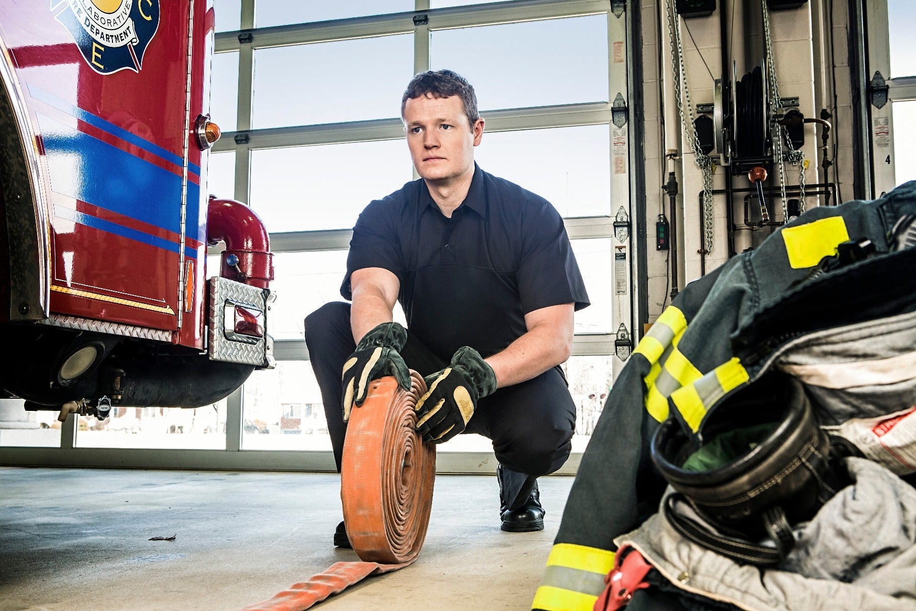Photograph of a firefighter in a fire station rolling a bright orange fire hose near a red firetruck with blue and gold stripes and a large bay door showing a residential area beyond a pile of firefighter gear with reflective yellow stripes and a dark helmet is visible