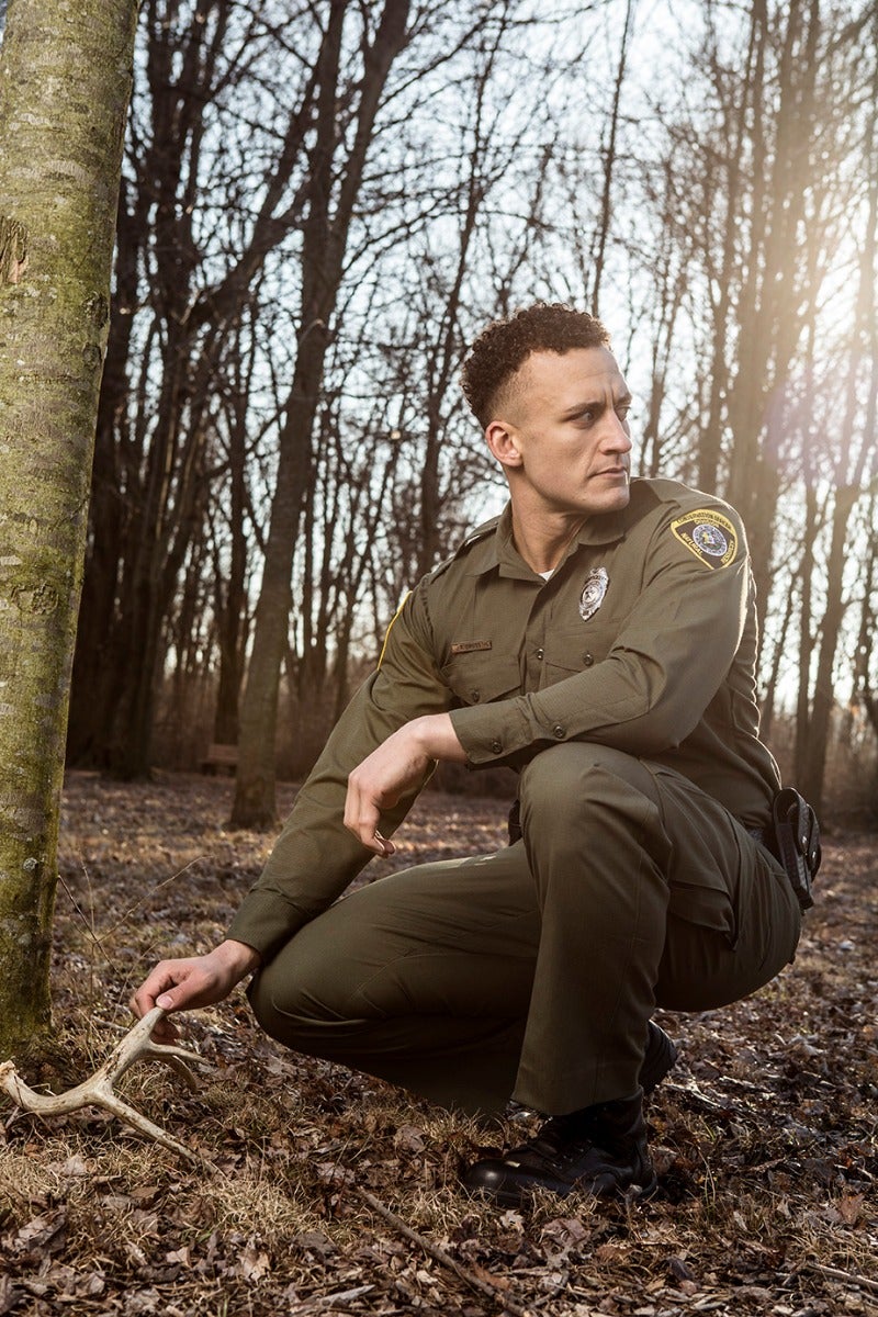 Photograph a park ranger in a forest examining a shed antler amongst bare trees and dry leaves his olive uniform and a sunlit background showcasing a badge and a hint of late afternoon light
