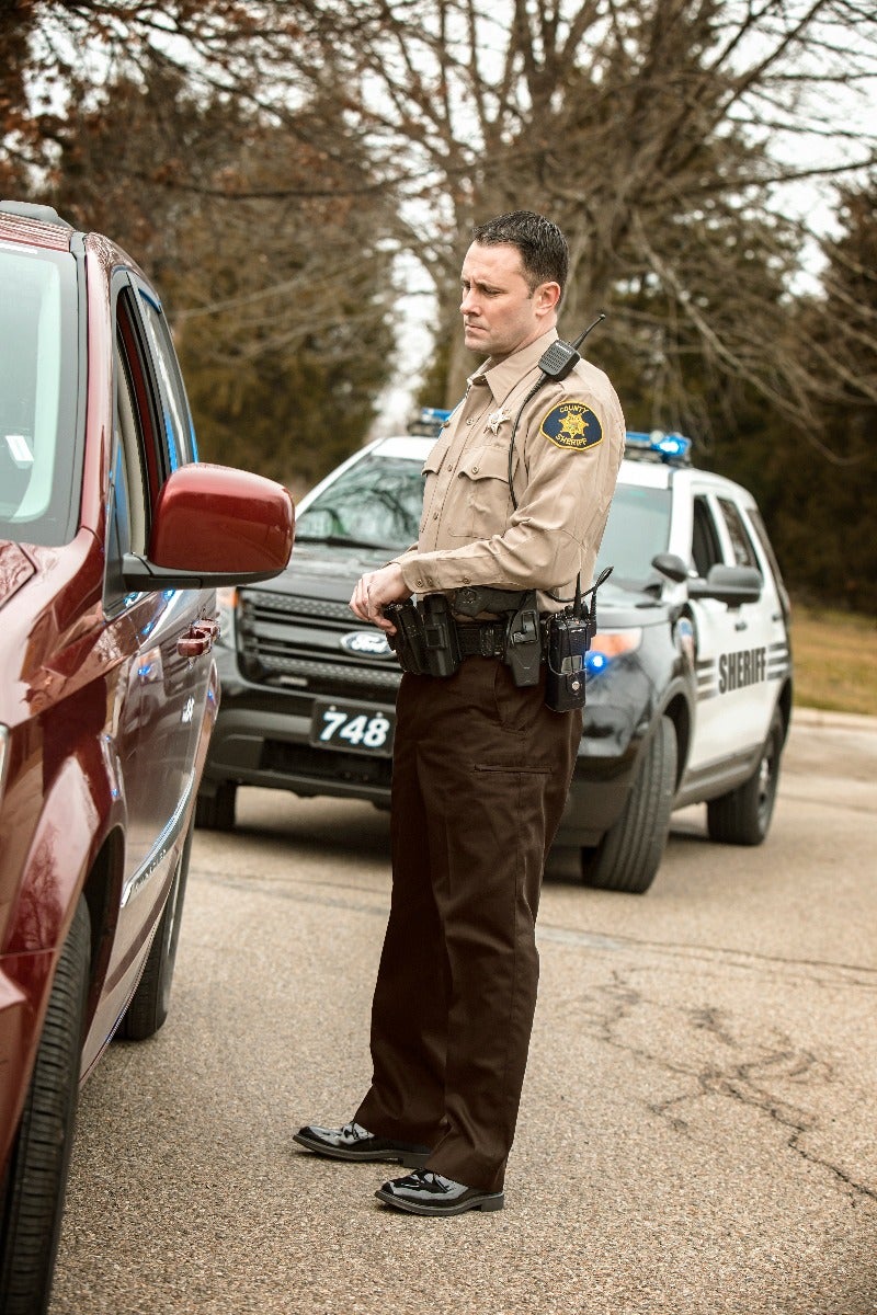 Photograph County Sheriff in brown uniform beside a maroon vehicle and a Sheriff's Department vehicle unit number 748 during a roadside stop with a black gun belt shiny shoes and a visible badge