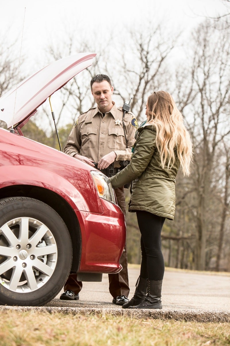 Photograph of a sheriff assisting a woman with her broken-down red car by the roadside amidst leafless trees showcasing a tan uniform, long blonde hair, and stylish black boots.
