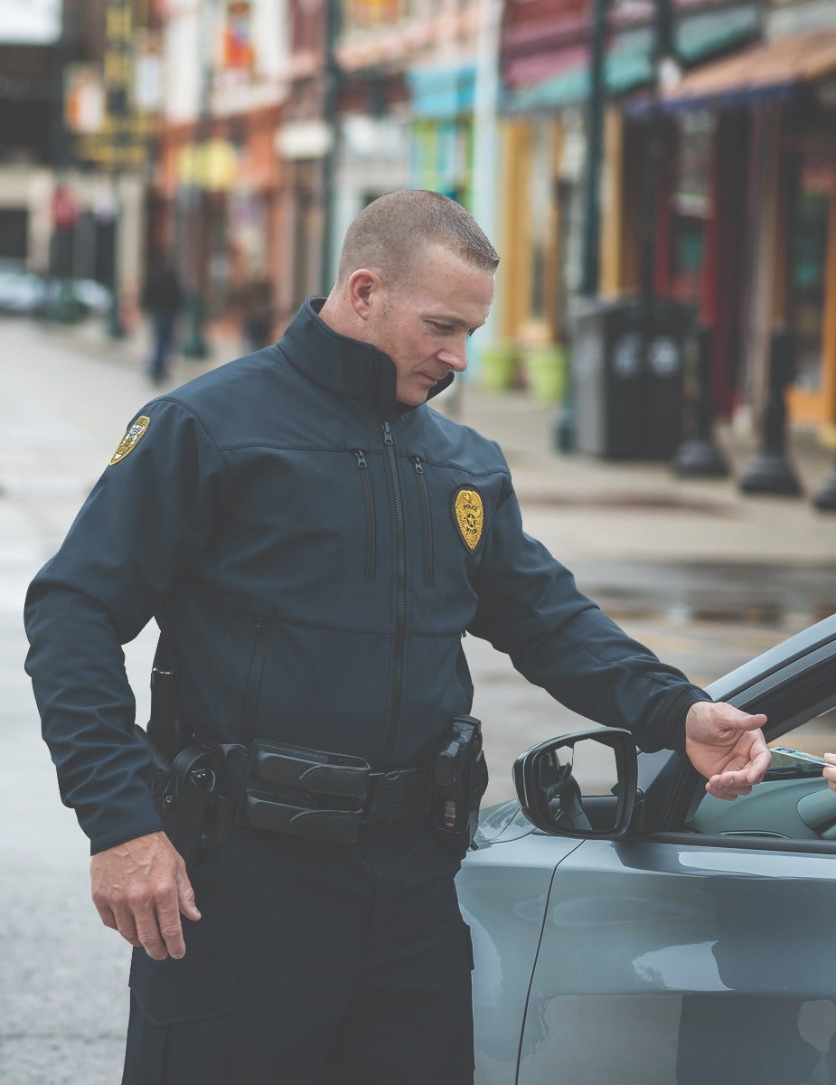 Photograph Police officer interacting with driver on a city street featuring colorful buildings and a grey car the officer wears a dark uniform with a gold badge and a black utility belt