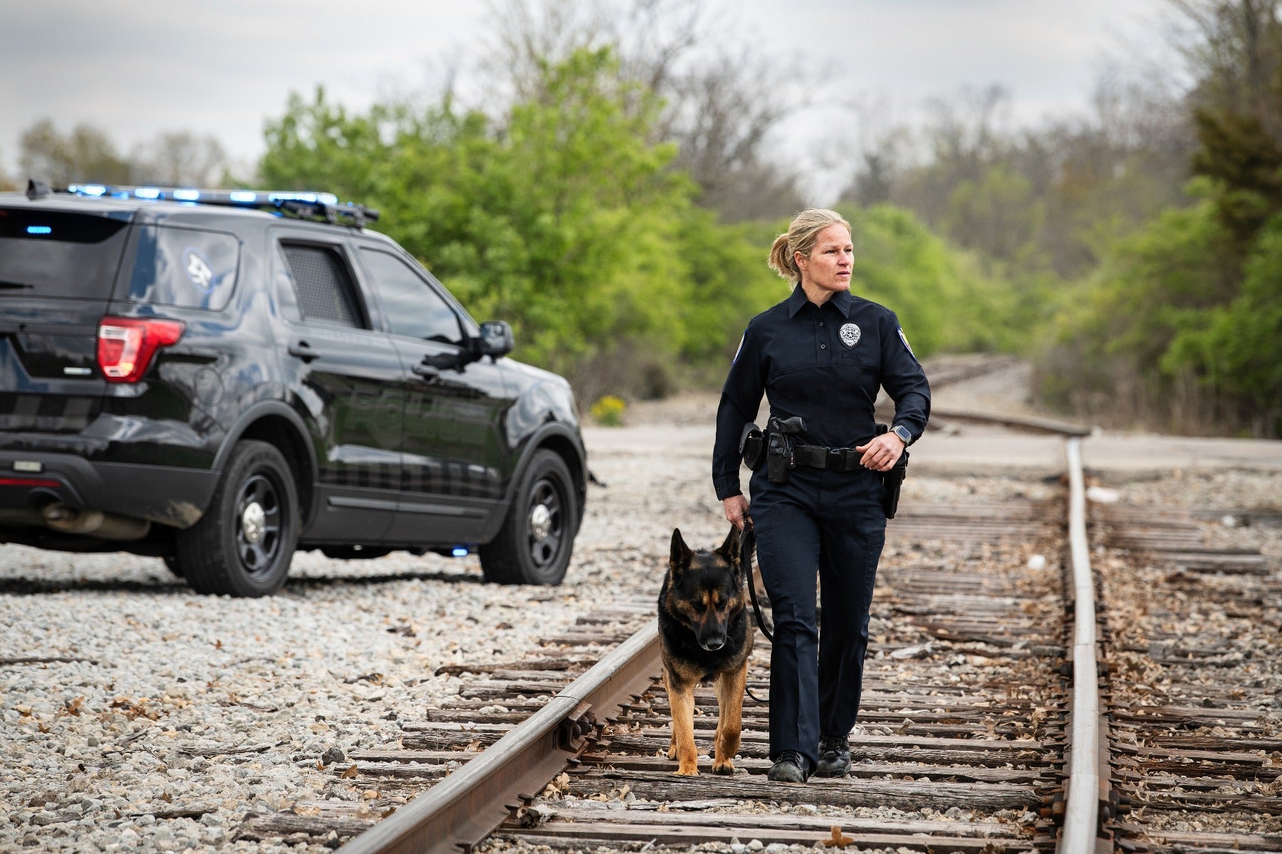 Photograph  A female police officer and her German Shepherd K9 partner walk along a rural railroad track near a black police SUV with flashing blue lights, amidst green trees and gravel