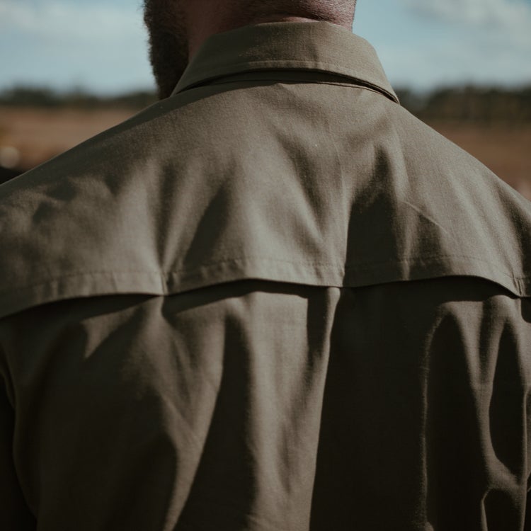 Photograph of a man's olive green shirt with a yoke detail and subtle texture showcasing the fabric's drape against a blurred outdoor background