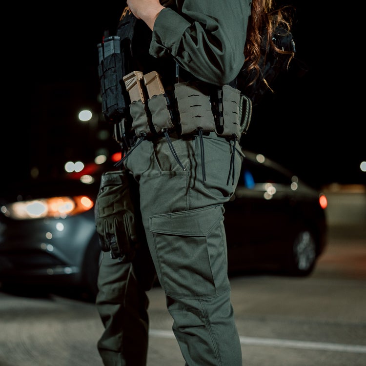 Photograph of a law enforcement officer in olive green tactical pants and duty belt with tan magazine pouches standing at night near a blurred dark car and city lights