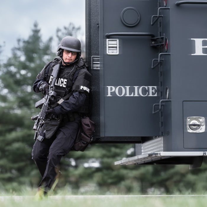 Photograph of a police officer in tactical gear leaning against a police armored vehicle outdoors the officer holds a rifle and wears a dark helmet and vest with POLICE insignia visible a dark bag is slung low on his left side