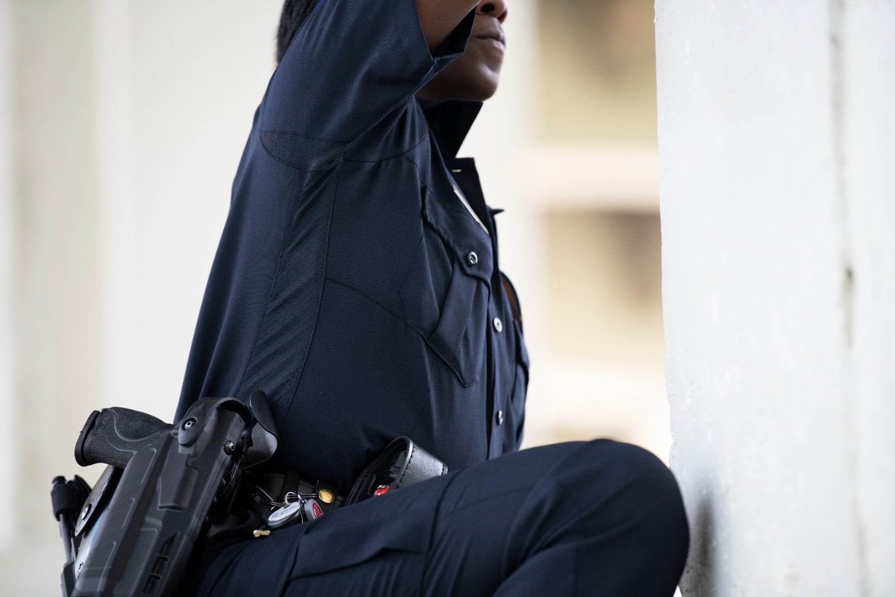 Photograph of a police officer in a dark blue uniform and duty belt near a white wall showcasing a firearm, badge, and keychain details.