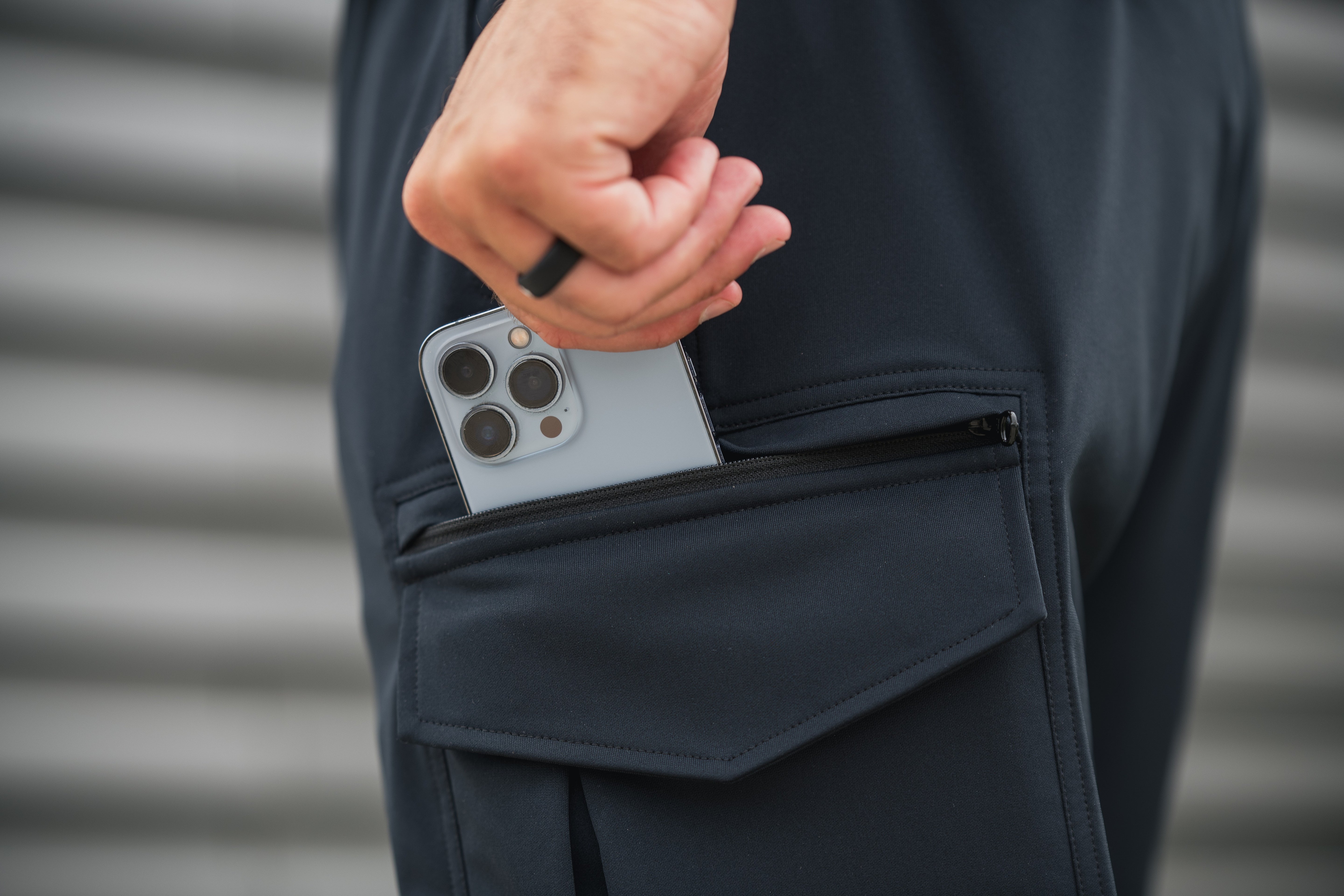 Photograph showing a light blue iPhone with a quad-camera system in a dark-colored pants pocket against a blurred grey corrugated metal background the phone has a sleek design and the pocket features a black zipper and subtle stitching