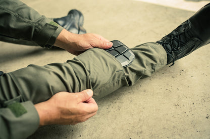 Photograph showing a person adjusting olive-green tactical pants with integrated black knee pads on a concrete floor while wearing black tactical boots the pants feature a Velcro patch and subtle stitching details
