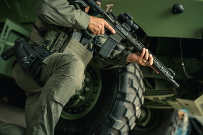 A photograph showing a soldier kneeling next to a military vehicle with a large tire and tactical gear including a rifle with scope and  a sidearm in a holster, featuring olive green tones and textured fabric details
