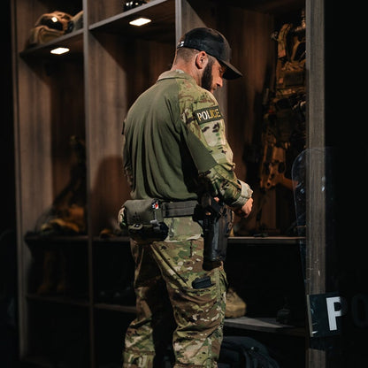 Photograph of a police officer in camouflage uniform and black hat inside an armory with shelves filled with weapons and tactical gear including a ballistic shield and a pistol holstered on his hip, showcasing his  duty belt and a dark green patch