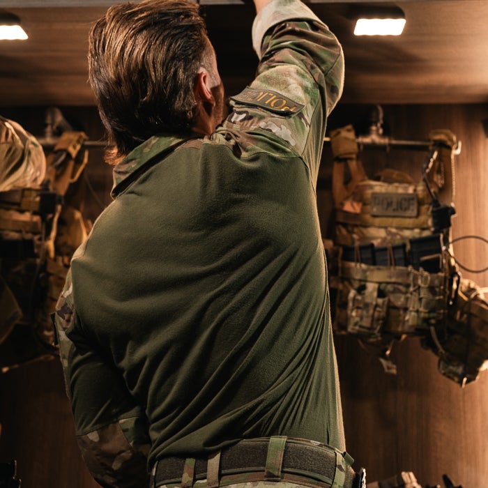 Photograph of a man in a camouflage uniform reaching for something in a dark room with tactical vests and subtle lighting showcasing the texture of his shirt and the Police patch on his arm
