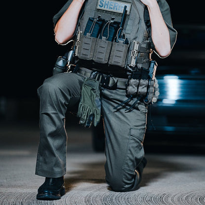 Photograph of a Sheriff kneeling at night near a blurred vehicle showing olive green tactical pants, vest with multiple pouches and a radio, and tactical gloves