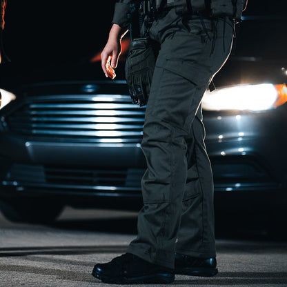 Photography Unknown Artist a police officer stands at night near a dark-colored car with bright headlights and a prominent grill wearing dark tactical pants and boots with dark gloves