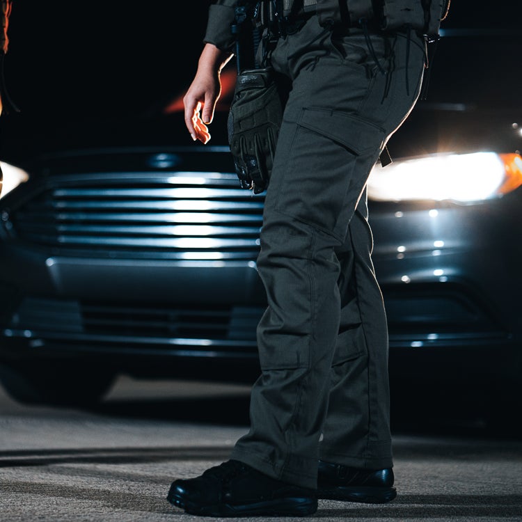Photography Unknown Artist a police officer stands at night near a dark-colored car with bright headlights and a prominent grill wearing dark tactical pants and boots with dark gloves