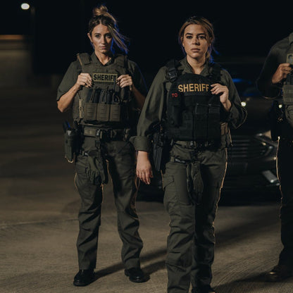 Photography  two female sheriff's deputies in tactical gear standing at night near a police vehicle with visible  "Sheriff" patches and utility pouches dark green uniforms and tactical belts
