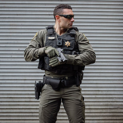 Photograph of a Sheriff's deputy holding a handgun against a corrugated metal backdrop wearing a tactical vest, olive green uniform, and protective eyewear.
