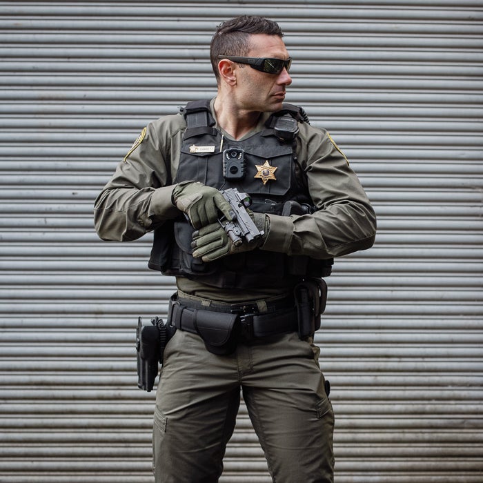 Photograph of a Sheriff's deputy holding a handgun against a corrugated metal backdrop wearing a tactical vest, olive green uniform, and protective eyewear.
