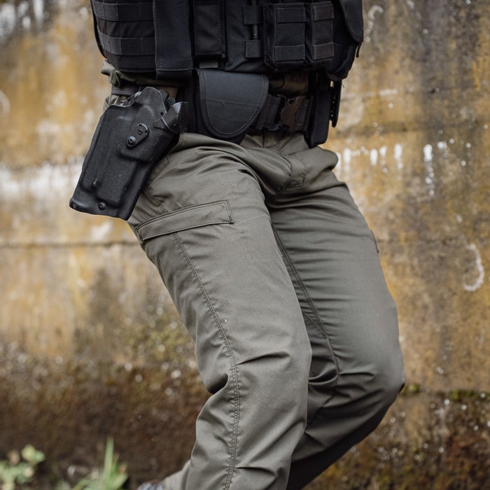 Photograph of a person wearing olive green tactical pants and a black ballistic vest with a handgun in a holster against a weathered stone wall showing detailed stitching cargo pockets and a sturdy belt