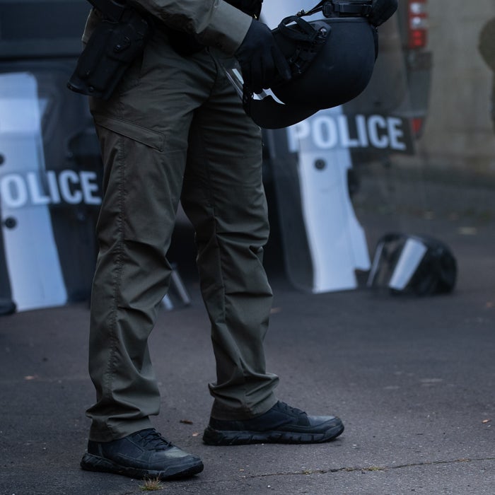 Photograph of a police officer in olive-green tactical pants and black boots holding a black helmet near police shields with the word POLICE visible in white lettering and featuring dark gray asphalt and a muted color palette